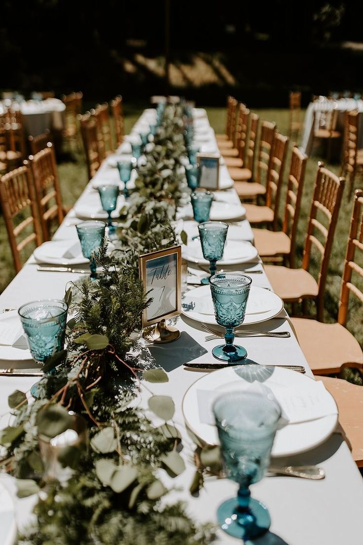 a long table is set with blue glassware and greenery for an outdoor wedding reception