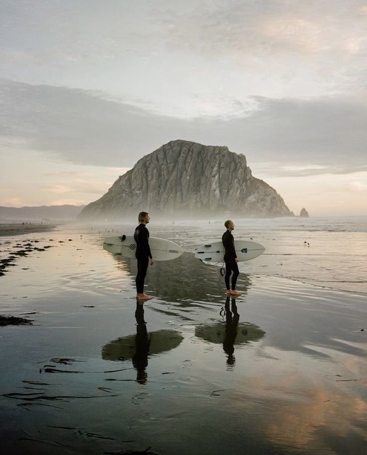 two people are standing on the beach in front of an island with a rock outcropping