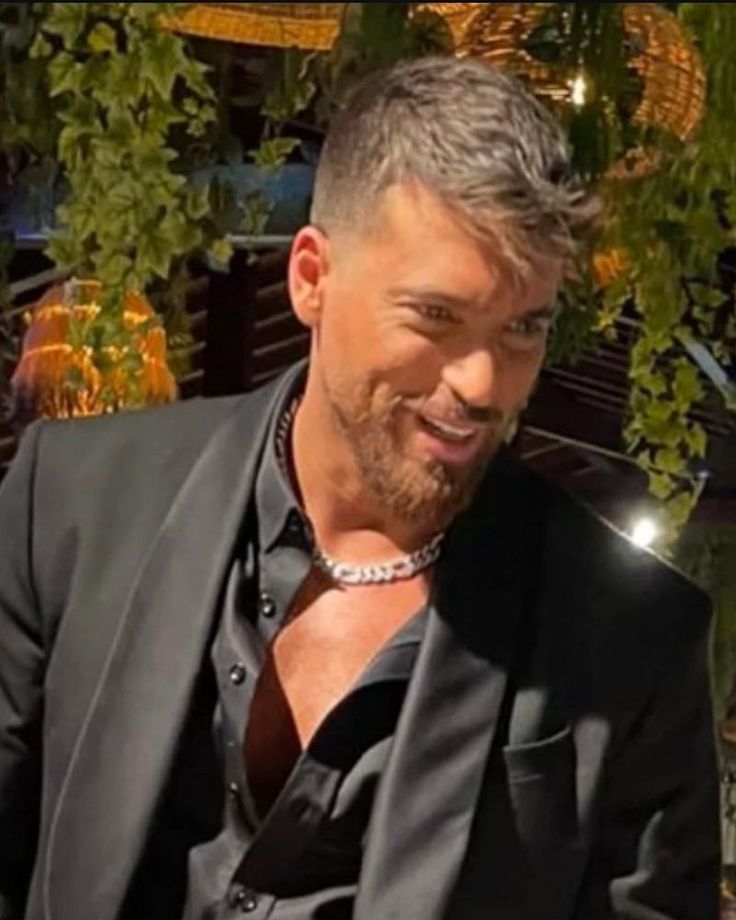 a man in a black suit and silver necklace smiling at the camera with his hand on his hip