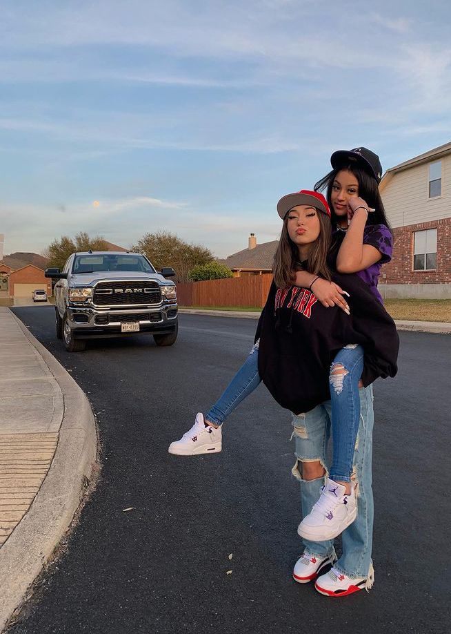 two girls standing in the middle of a street with their arms around each other while one girl is on her cell phone