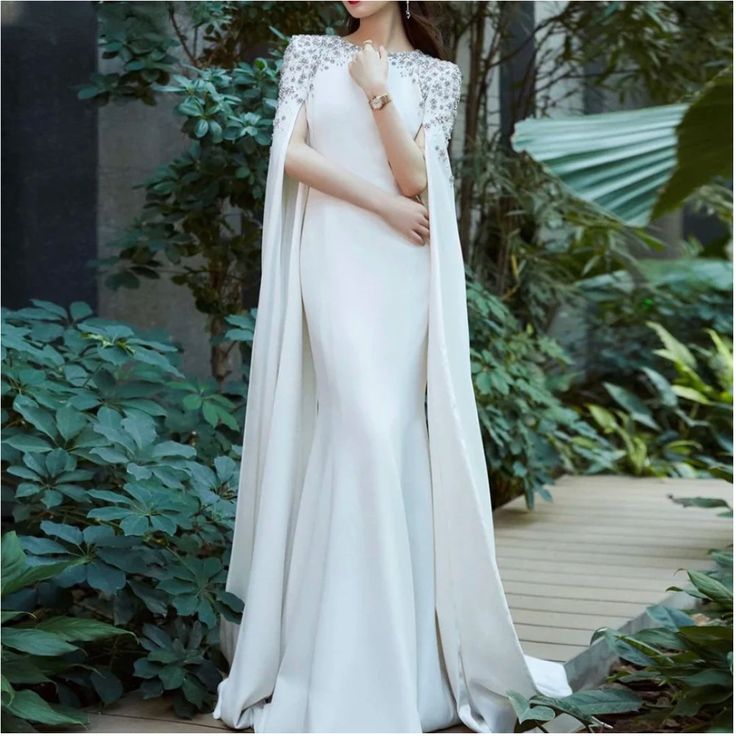 a woman in a white gown and cape standing on a walkway with greenery behind her