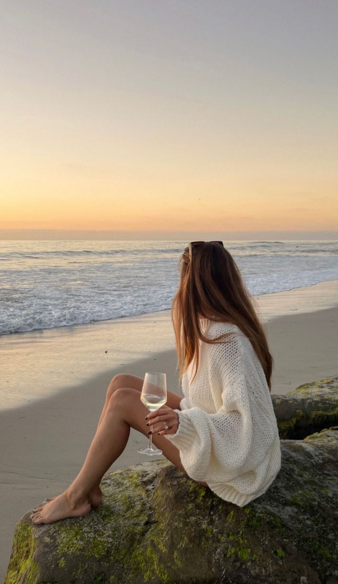 a woman sitting on top of a rock next to the ocean holding a glass of wine