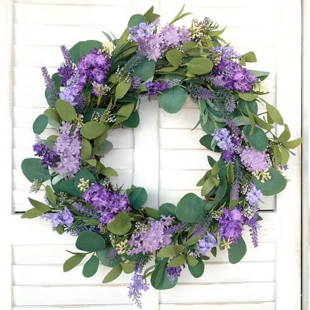 a wreath with purple flowers and greenery is hanging on a white brick wall in front of a door