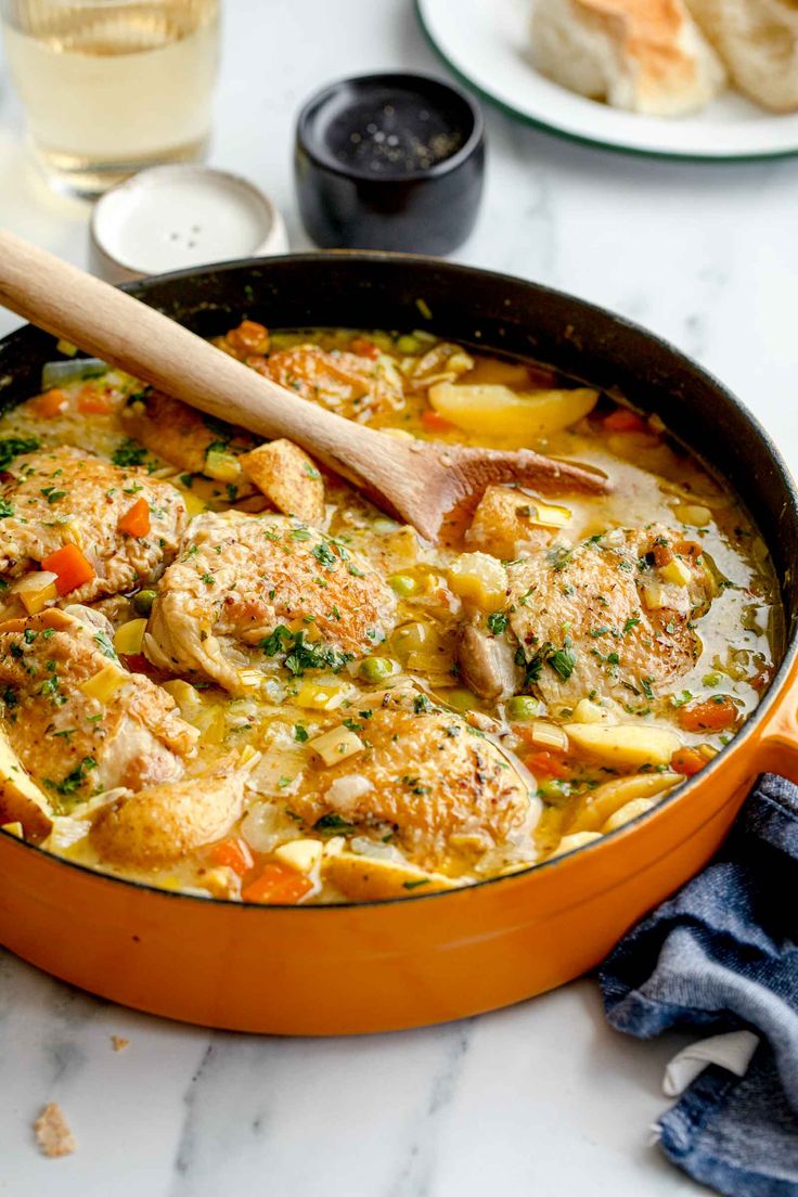 an orange pan filled with chicken and vegetable stew next to bread on a white table