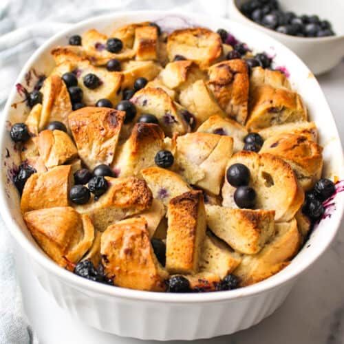 blueberry bread pudding in a white dish with fresh blueberries and other toppings