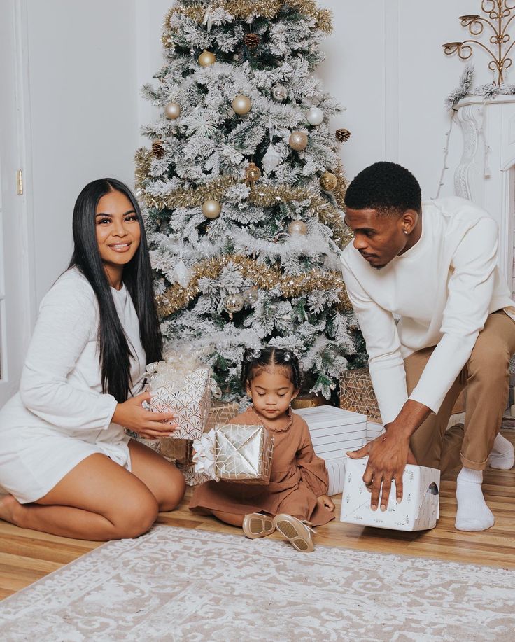 a man and woman opening presents next to a christmas tree with a small child sitting on the floor