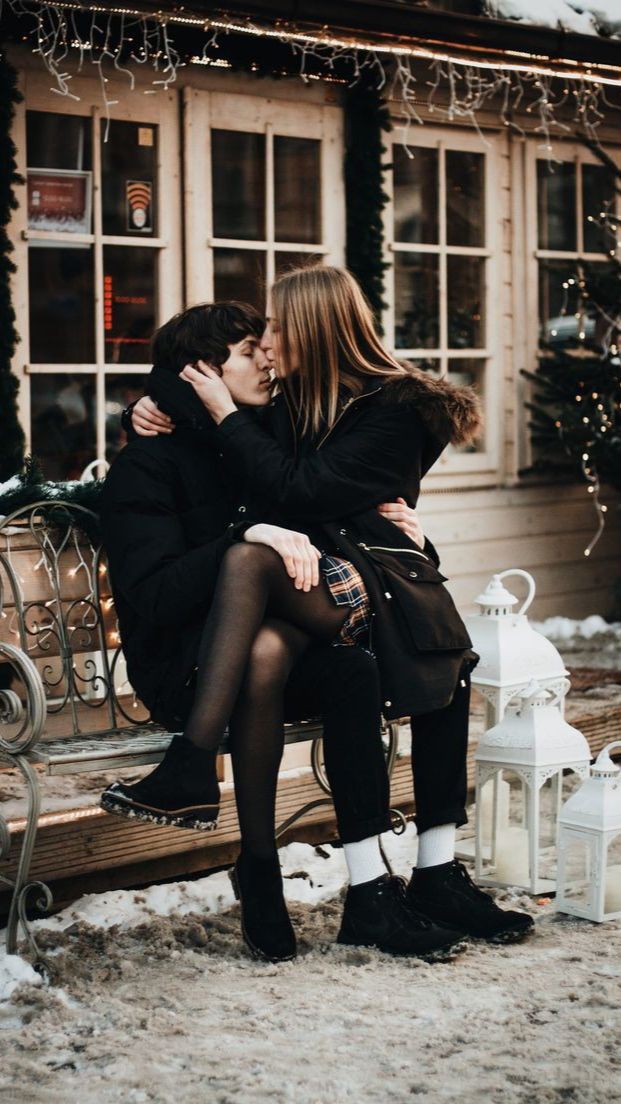 two people sitting on a bench in front of a building with snow and christmas lights