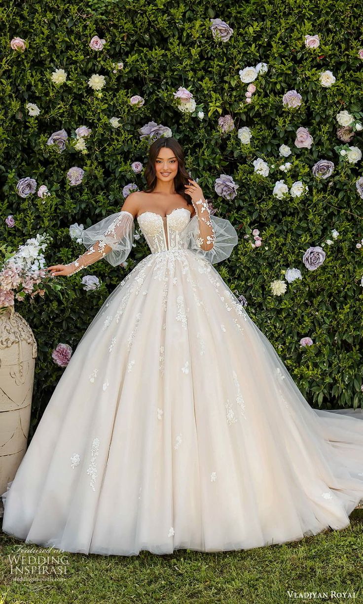 a woman standing in front of a hedge wearing a wedding dress with long sleeves and flowers on it