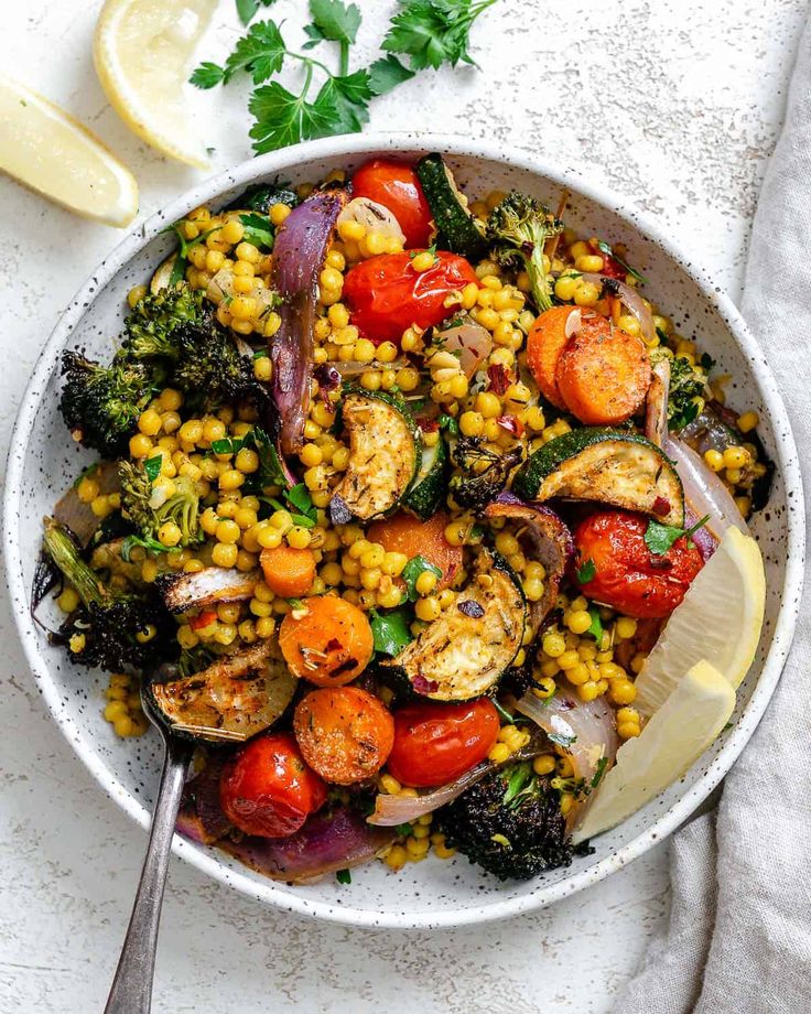 a white bowl filled with corn, tomatoes and broccoli next to lemon wedges