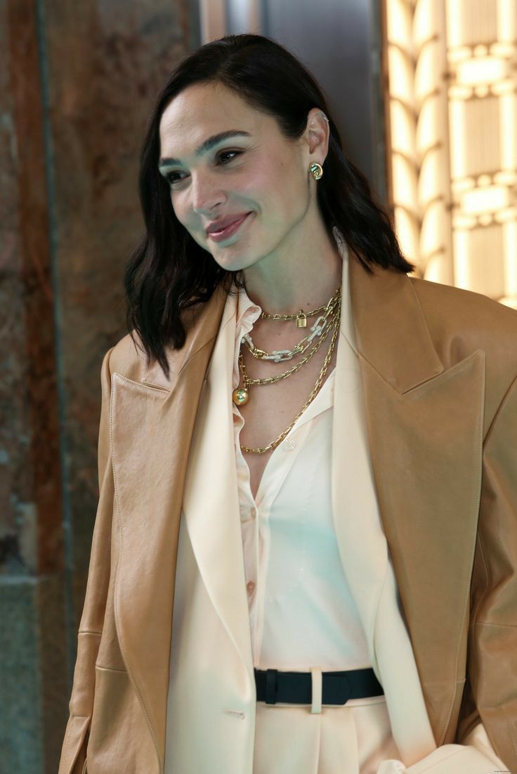 a woman wearing a tan jacket and white shirt with gold jewelry on her neck, standing in front of a wall