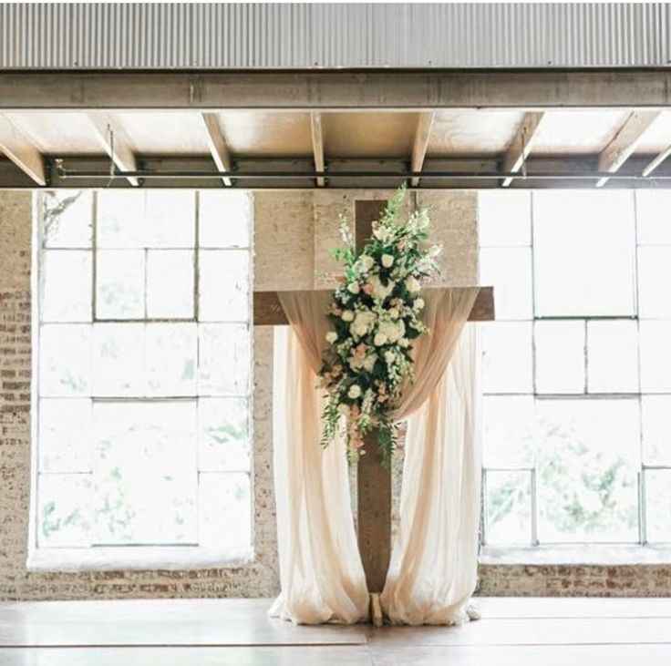 a cross decorated with flowers and greenery in front of large windows at an indoor wedding venue