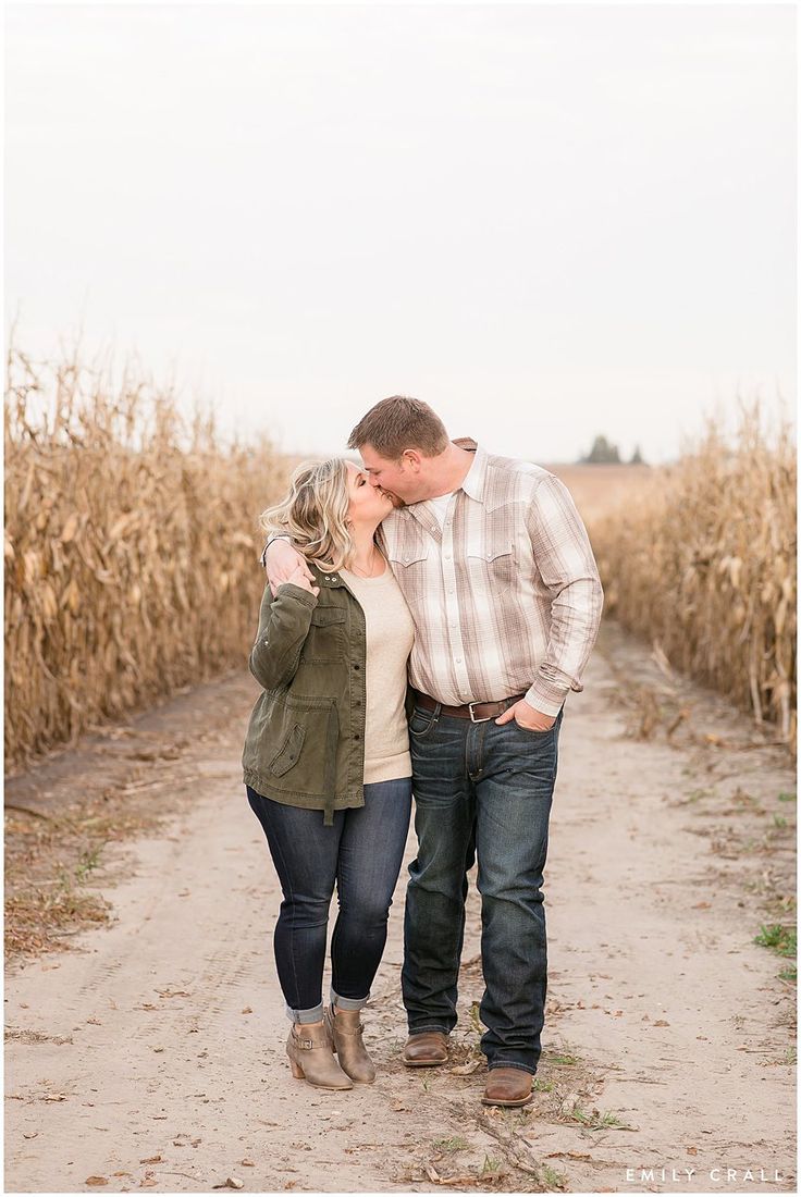 a man and woman walking down a dirt road in front of a cornfield during their engagement photo session