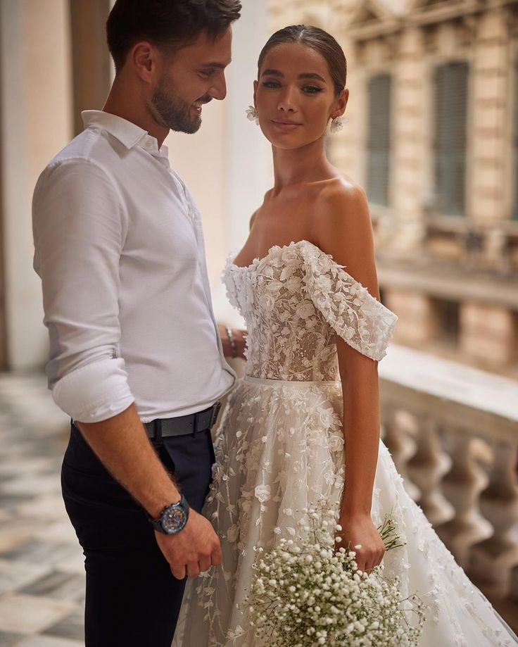 a man standing next to a woman in a wedding dress and holding a flower bouquet