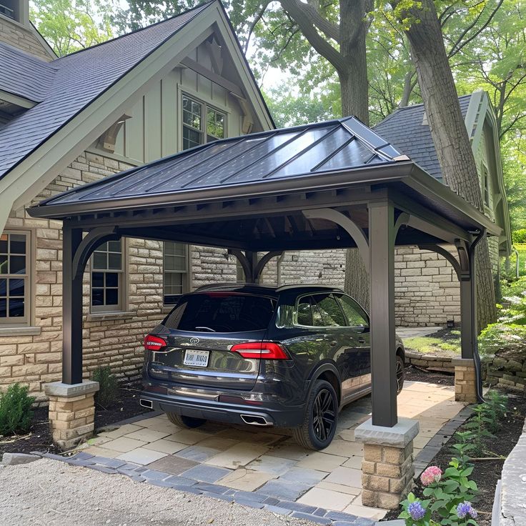 Elegant carport featuring a dark gray metal roof, beautifully complemented by tan stone accents and limestone piers. This open-sided structure shelters a black Volkswagen Tiguan, set against an old cottage-style house with black windows. The landscaping boasts lush greenery, colorful flowers, and two large decorative brown blocks enhancing the aesthetic. Carport Beside House, Beautiful Carport Ideas, Carport Driveway Ideas, Unattached Carport Ideas, Pergola As Carport, Cottage Carport Ideas, Adding A Carport To Front Of House, Carport With Privacy Wall, Detached Carport Ideas Breezeway