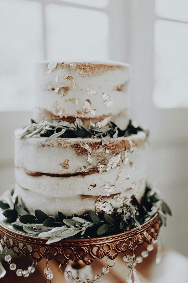 a three tiered wedding cake with greenery on the top and sides, sitting on a gold platter