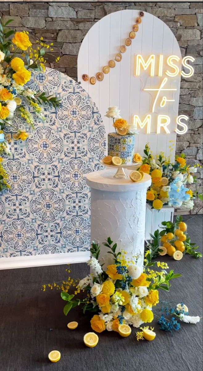 a white cake with yellow and blue flowers on it sitting in front of a sign that says miss 75 mr's