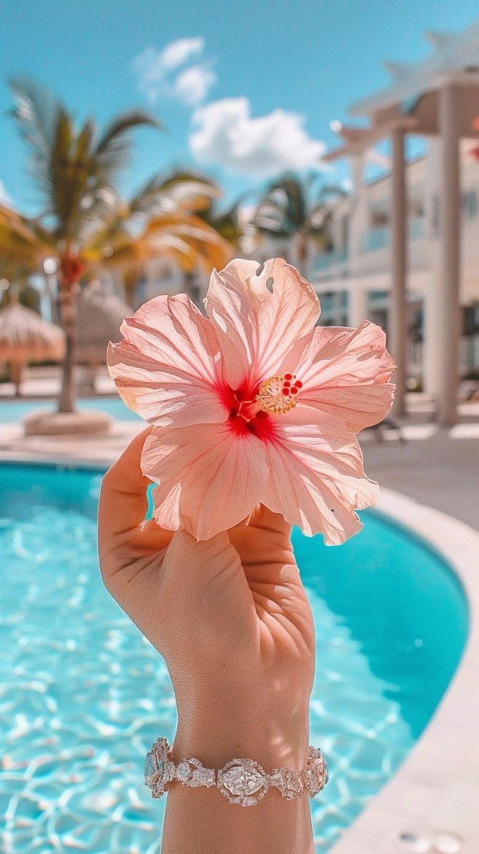 a person holding up a pink flower in front of a swimming pool with palm trees