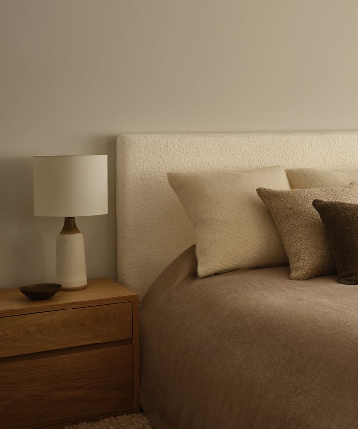 a bed with three pillows on top of it next to a night stand and lamp