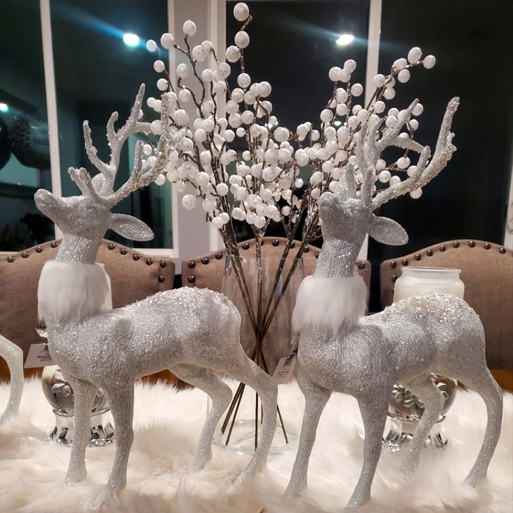 two silver reindeer figurines sitting on top of a white fur covered floor next to a vase filled with flowers