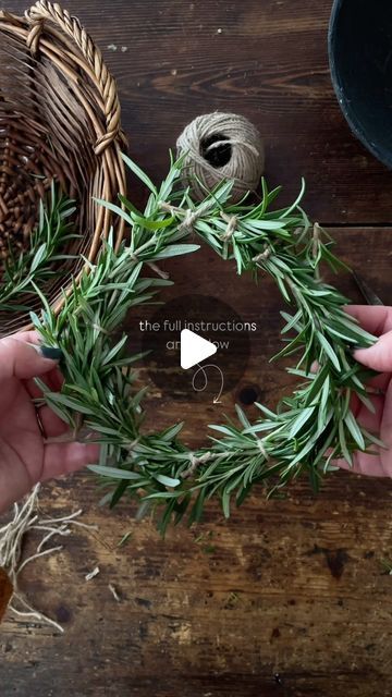 two hands are holding a wreath made out of green leaves and twine on top of a wooden table