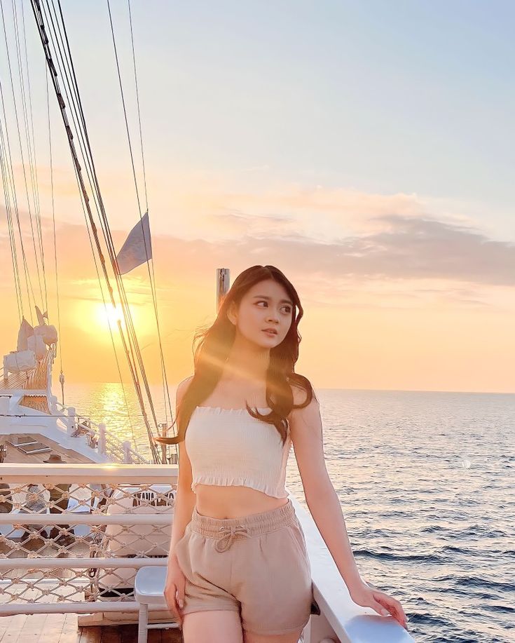 a woman standing on the deck of a boat at sunset with her hands in her pockets