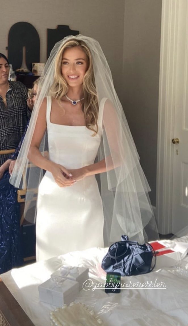 a woman standing in front of a bed wearing a white wedding dress and holding a veil over her head