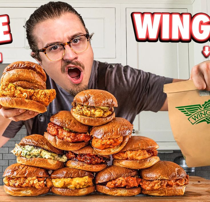 a man holding up a large sandwich in front of a pile of sandwiches on a table