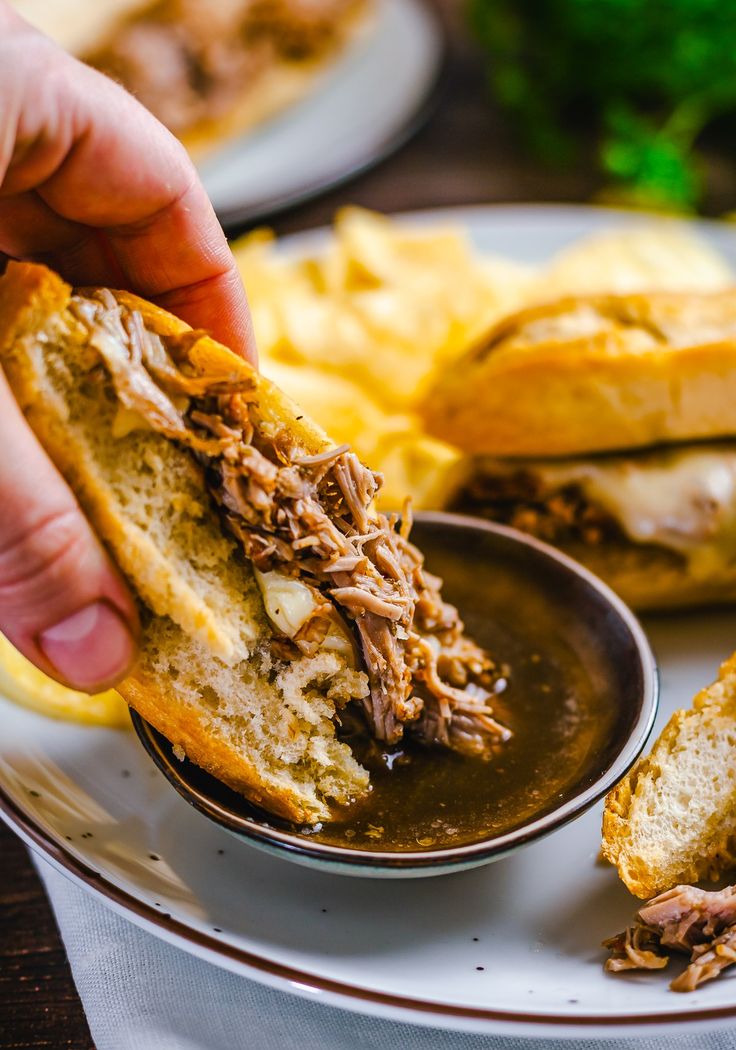 a person holding a sandwich over a plate with other food on the table behind it