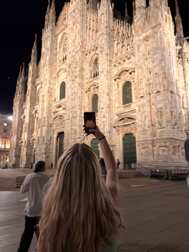 a woman holding up her cell phone in front of a cathedral at night with people walking by