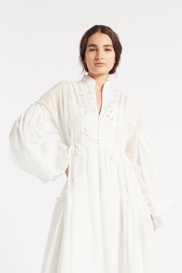 The JUNO dress with gathered full sleeve detail and skirt panels has a high low hem that creates an elegant silhouette. Seen here in Soft White cotton silk voile with floral silk embroidered bib front detail trimmed with pearl buttons. Long Sleeve Dresses With Gathered Sleeves For Garden Party, White Dresses With Blouson Sleeves, Spring Daywear Cotton Voile Dress, Spring Cotton Voile Day Dress, Silk Flowy Dress With Sheer Sleeves, Spring Cotton Dresses For Daywear, Flowy Silk Dress With Sheer Sleeves, Billowy Blouson Sleeve Dresses For Daywear, Silk Dress With Gathered Long Sleeves