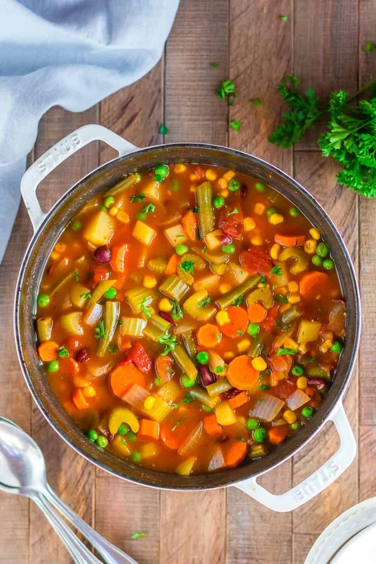 a pot filled with vegetable soup on top of a wooden table next to spoons