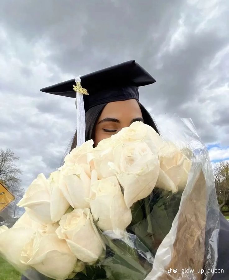 a woman wearing a graduation cap and gown holding flowers in front of her face with the sky behind her