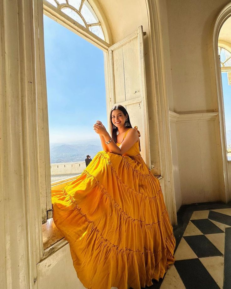 a woman in a long yellow dress standing by an open window looking at the camera