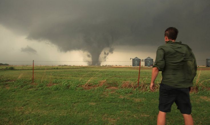 a man standing in the grass looking at a large tornado coming from behind his house