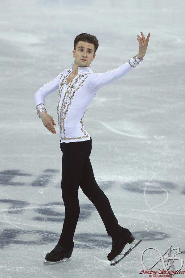 a male figure skating on the ice in a white shirt and black pants with his arms outstretched