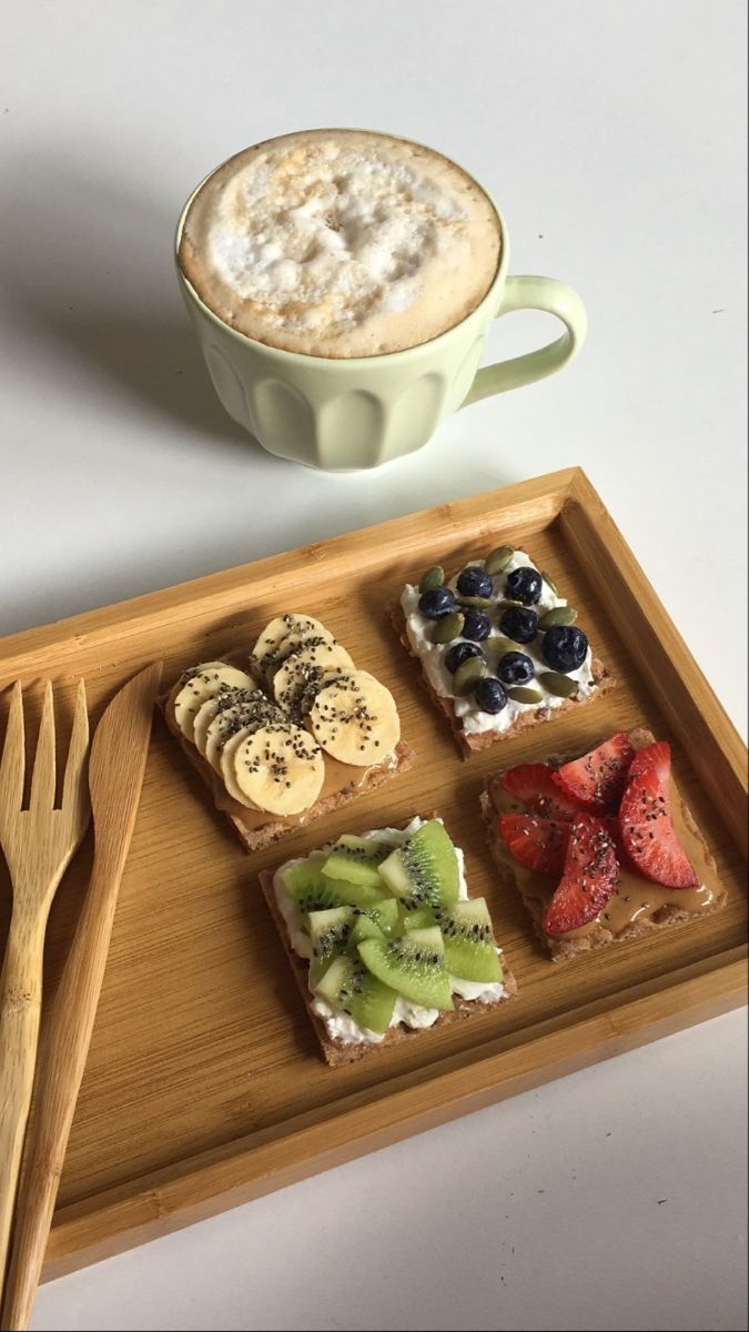 a wooden tray topped with different types of food next to a cup of cappuccino