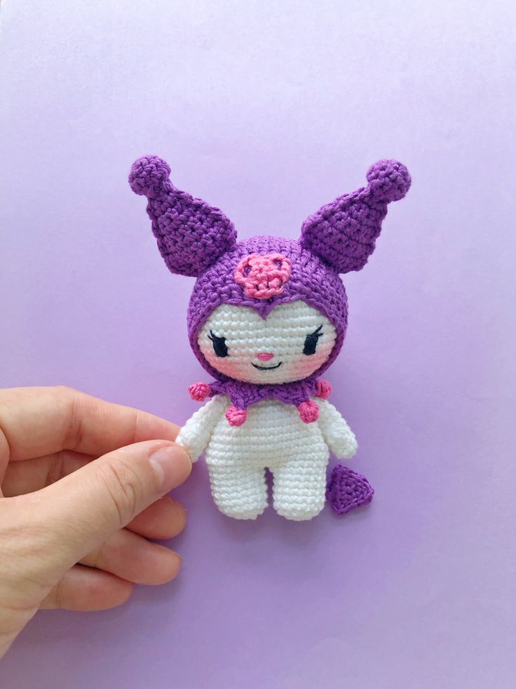 a hand is holding a small crocheted bunny doll on a purple background with pink flowers