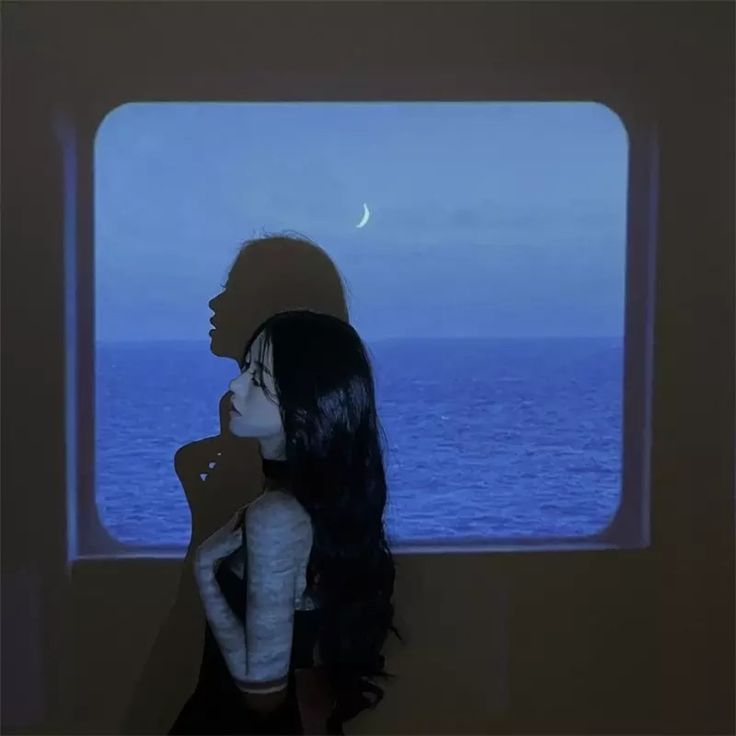 a woman standing in front of a window looking out at the ocean with a half moon behind her