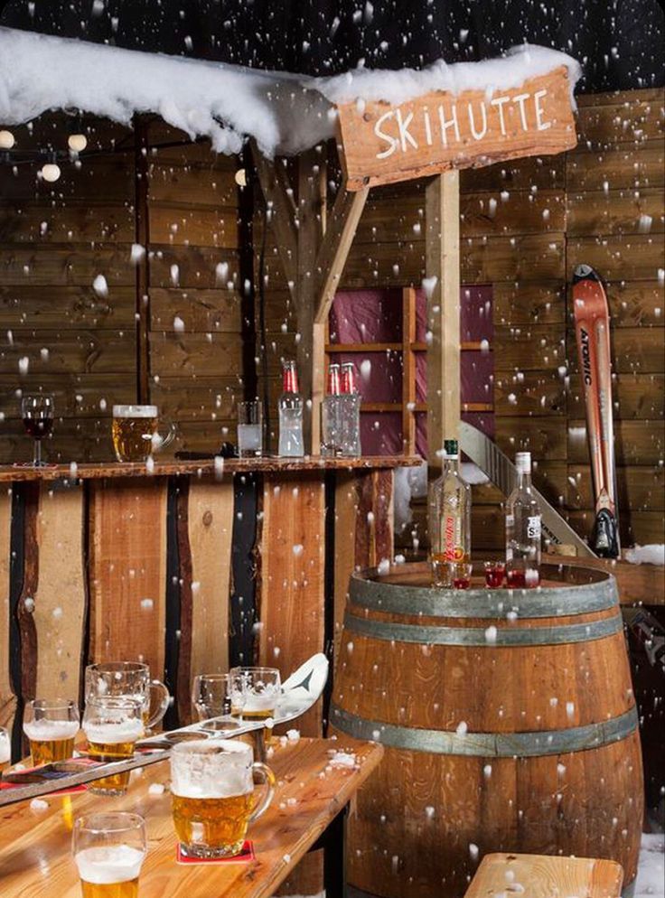 a wooden table topped with lots of glasses filled with beer next to barrels covered in snow