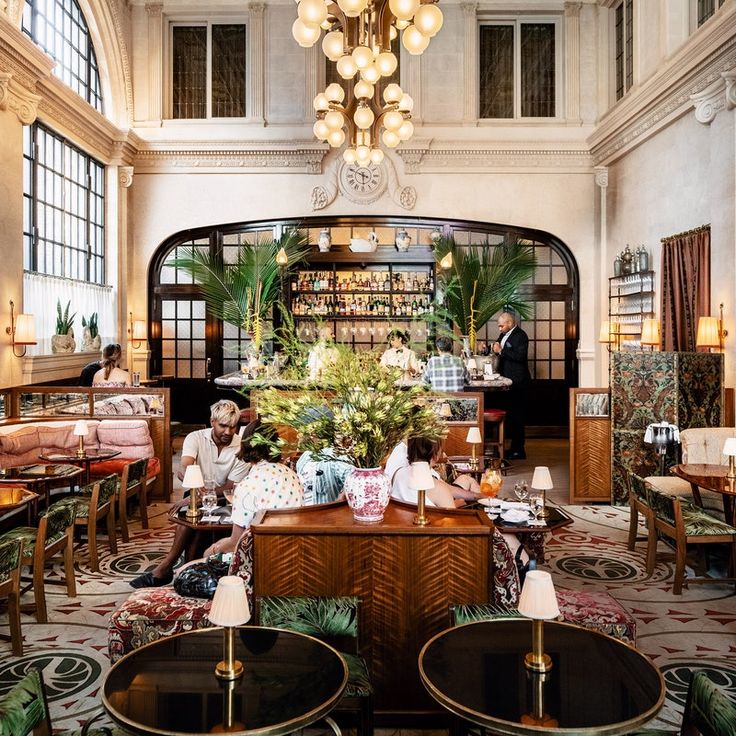 people sitting at tables in a large room with chandeliers and plants hanging from the ceiling