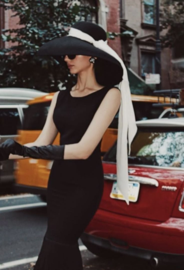 a woman in a black dress and hat is walking down the street with her hand on her hip