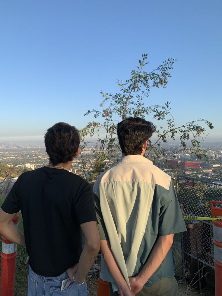 two young men standing next to each other on top of a hill looking at the city