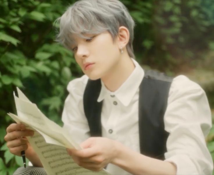 a person sitting down reading a book in front of some trees and bushes with their eyes closed