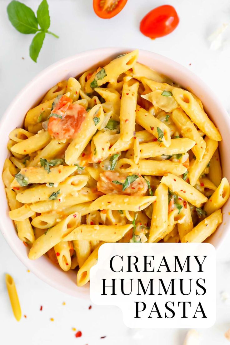 creamy hummus pasta in a white bowl with tomatoes and basil on the side text overlay reads creamy hummus pasta