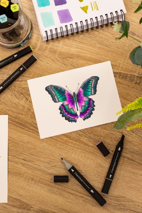 some markers and pens are on a table next to an open notebook with a butterfly drawn on it