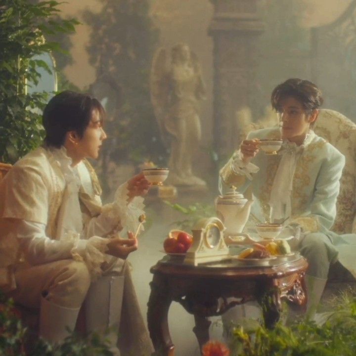 two people sitting on chairs drinking tea in front of a table with fruit and flowers