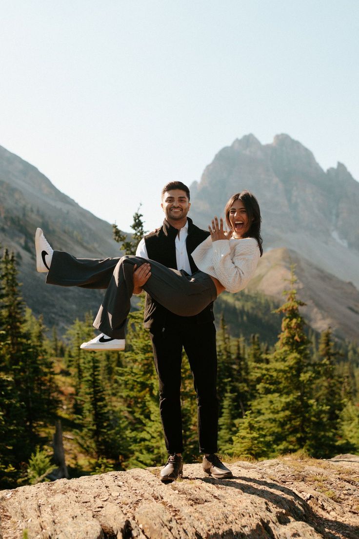 a man carrying a woman on his back while standing on top of a rock in the mountains