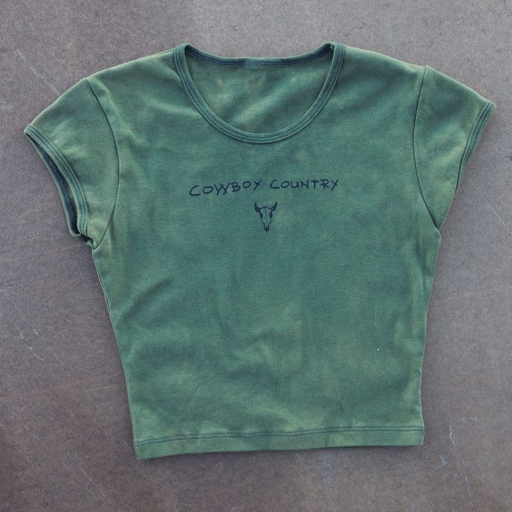 Our Cowboy Country tee is printed on a forest green women’s L.A. Apparel cap sleeve baby rib crop top tee with dark green ink. This baby tee features a flattering modest crop and pairs perfectly with jean shorts or sweats. Montana is cowboy country, where the rivers run with tales of cattle drives and the rugged landscapes bear the hoof prints of legends. Pay homage to this sentiment with this tee; ready to be worn to a country dance, on a hike or while working in your garden! The designs are printed on the front center of the shirt and also on the center back. Design by Paige Johnson in Bozeman, Montana. Made in U.S.A 1x1 Baby Rib 100% Combed Cotton Fitted Graphic Print T-shirt For Ranch, Fitted Casual T-shirt For Outdoor, Casual Fitted T-shirt For Outdoor, Western Style Tops For Country Concerts And Festivals, Western Cotton Tops For Country Festivals, Fitted Crew Neck Top For Rodeo, Casual Tops For Rodeo And Country Festivals, Graphic Print Crew Neck Tops For Country Festivals, Fitted Graphic Print Tops For Country Concerts