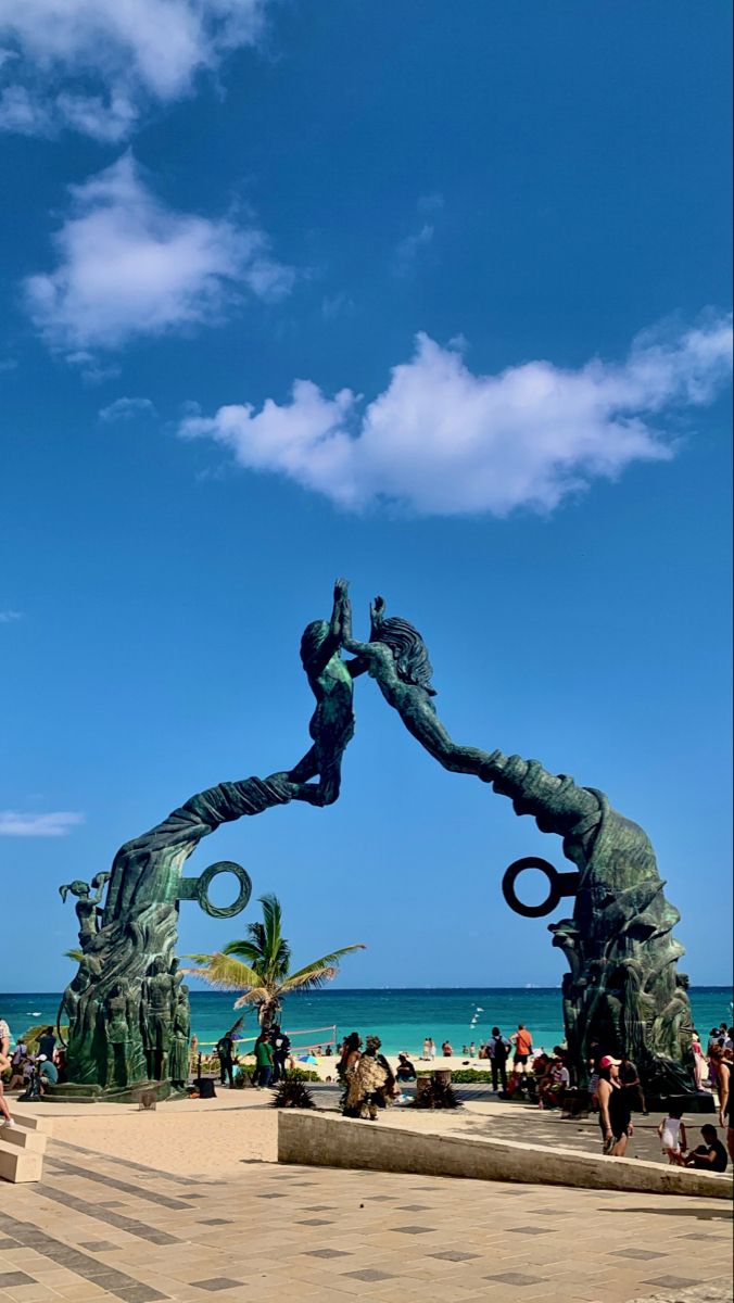 people are sitting on benches near the water and statues in front of the beach with blue sky