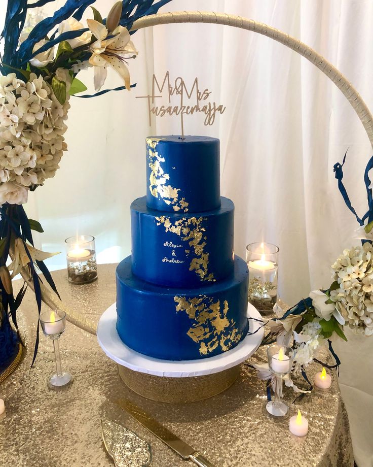 a blue wedding cake with gold leaf designs on the top and bottom, surrounded by candles